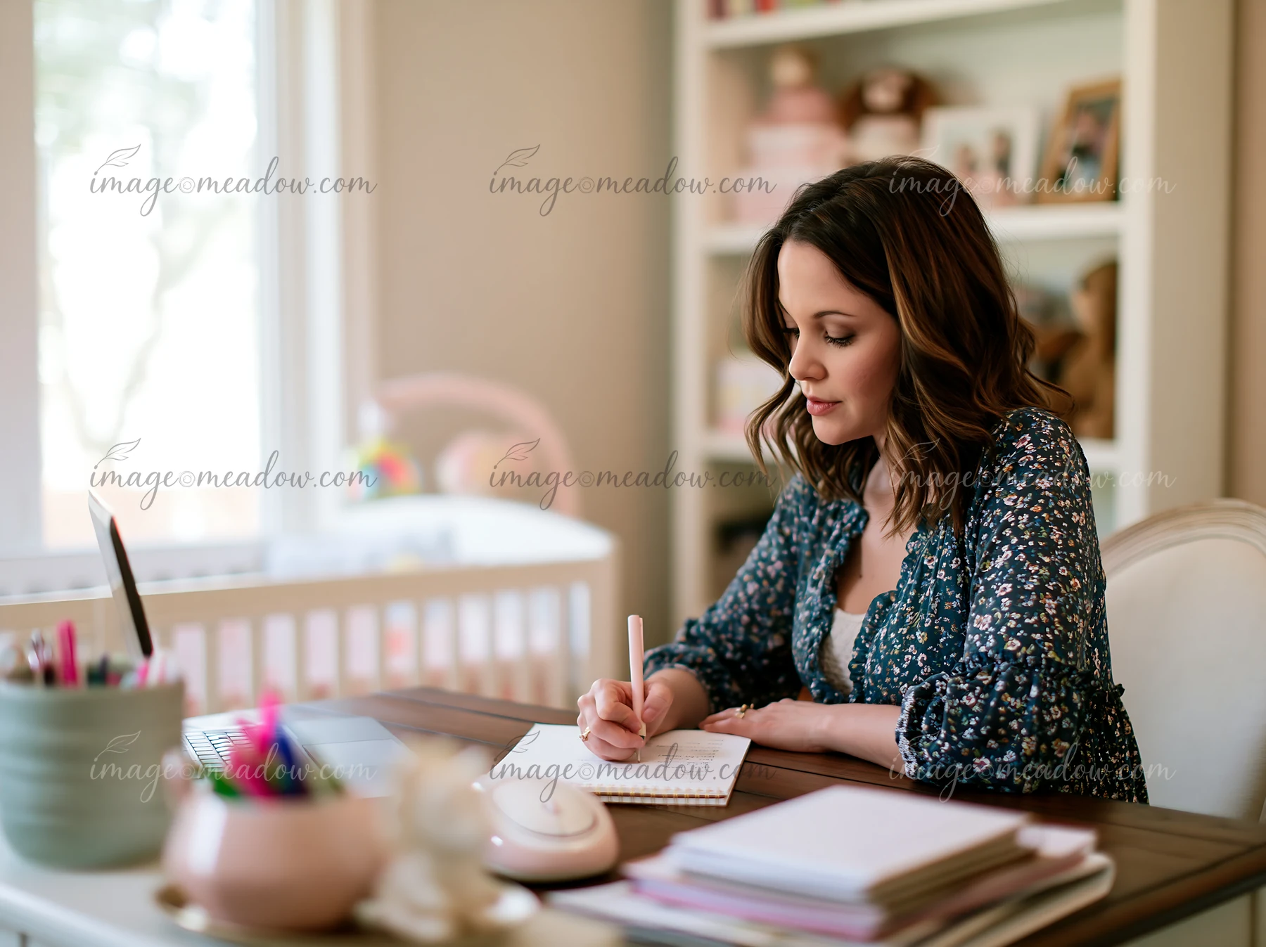 Woman writes in a notebook at a desk in a bright home workspace beside a laptop and books.