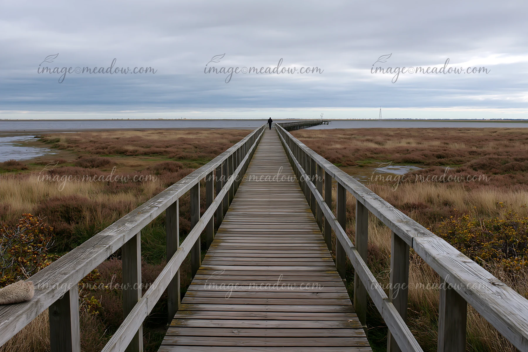 Wooden boardwalk crosses a salt marsh toward calm coastal water beneath a cloudy sky, with one person ahead.