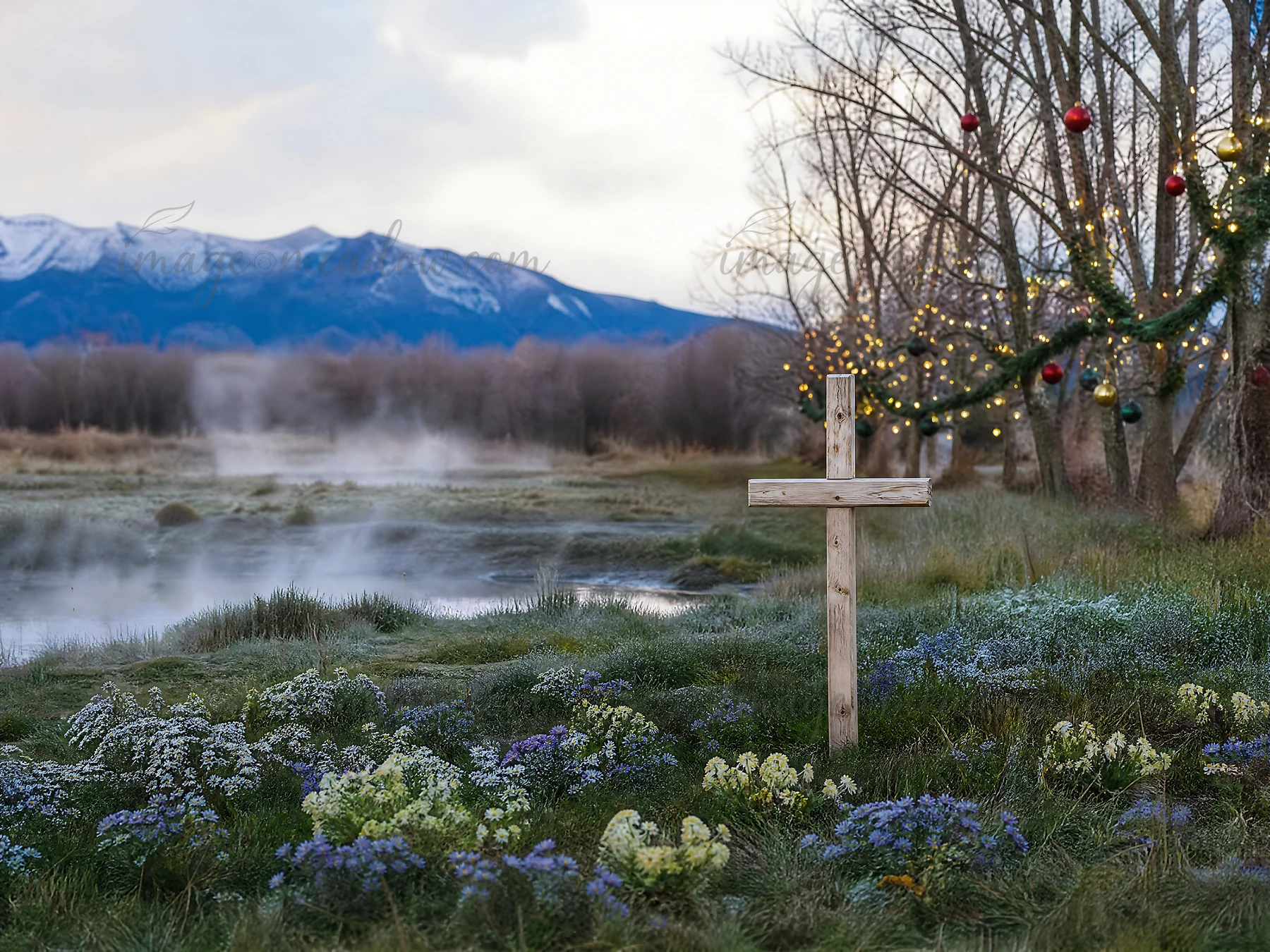 A wooden Christian cross stands in a meadow near hot springs, surrounded by flowers and trees. Snow-capped mountains are visible in the background under a cloud