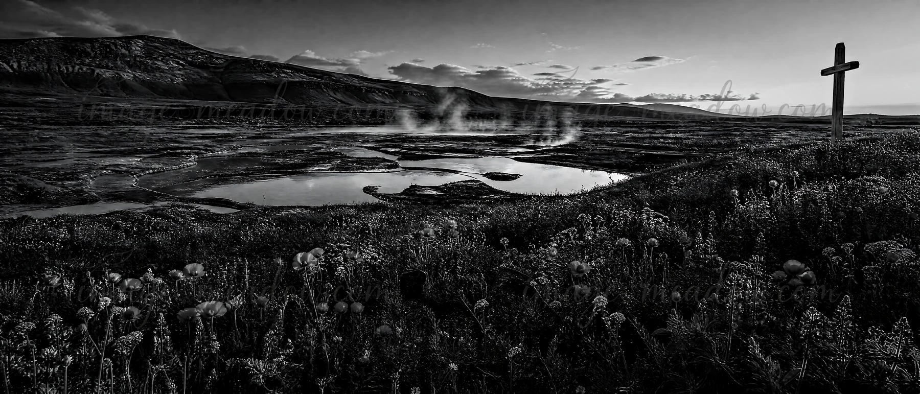 A wooden Christian cross stands in a meadow by hot springs. Steam rises from the ground, and the landscape features mountains and scattered vegetation. This sce