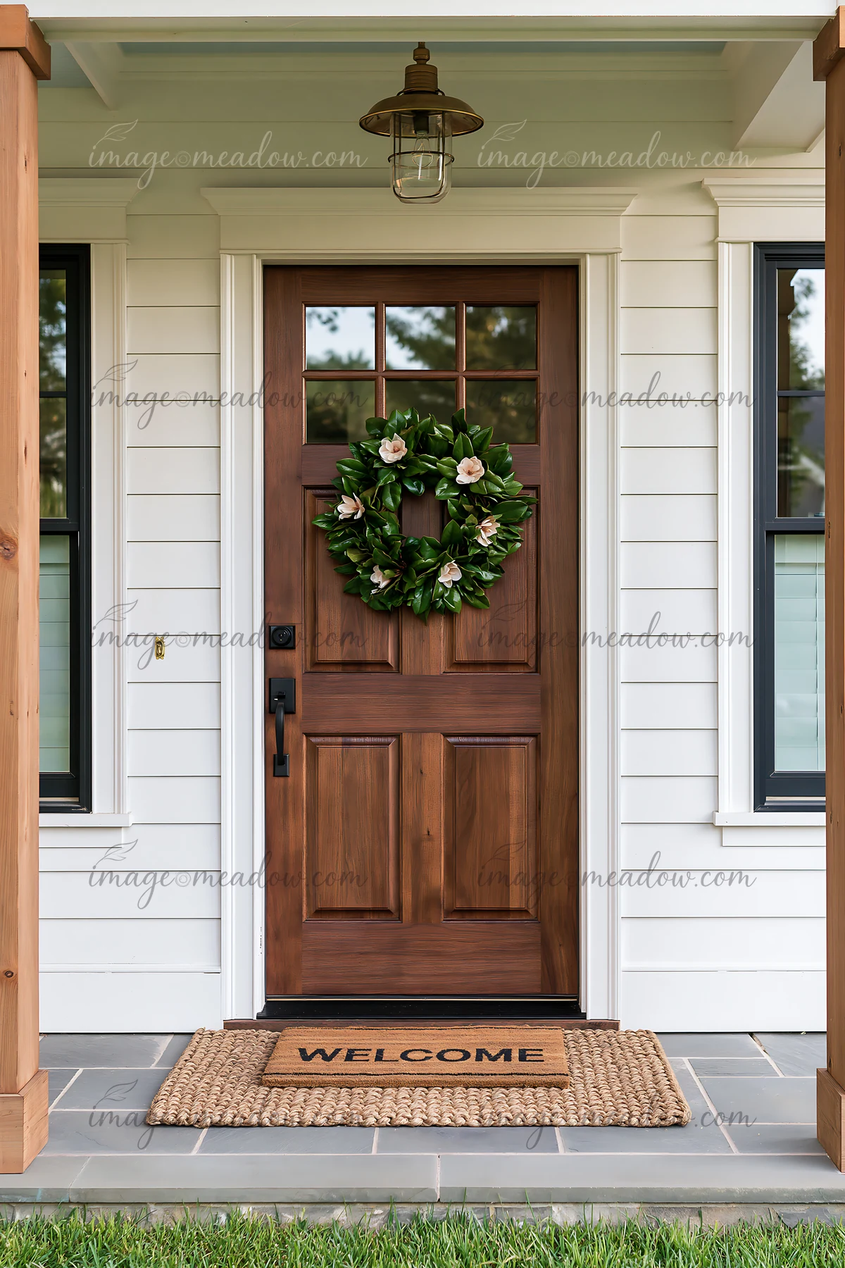 Wooden entrance door with a leafy floral wreath and welcome mat beneath a covered porch lantern.