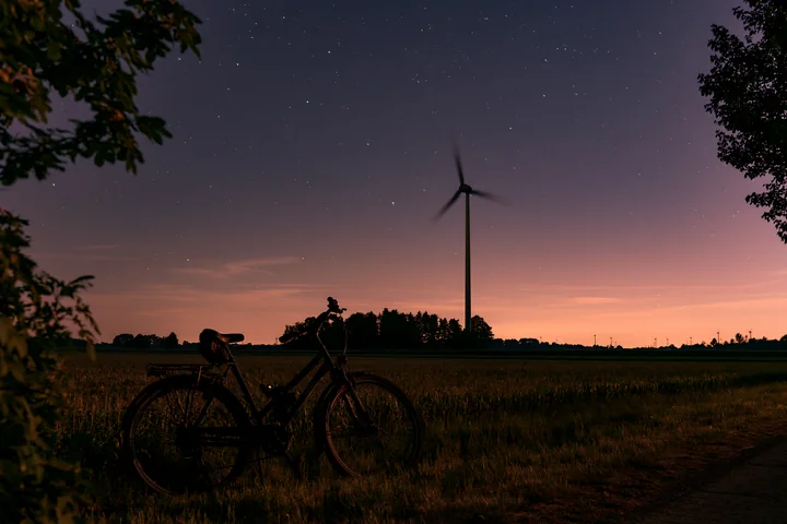 A bicycle stands near a wind turbine in a rural field under a starry sky.