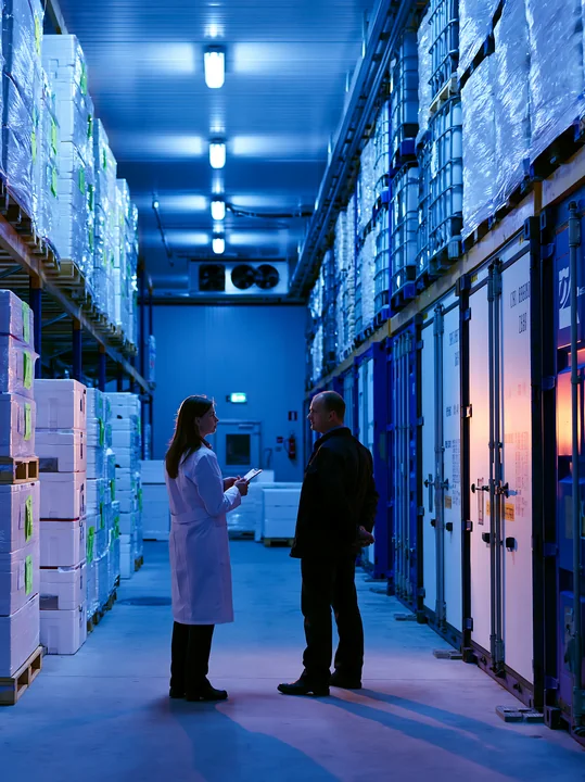 A businesswoman discusses details with a supplier in a warehouse aisle filled with stacked boxes and crates.