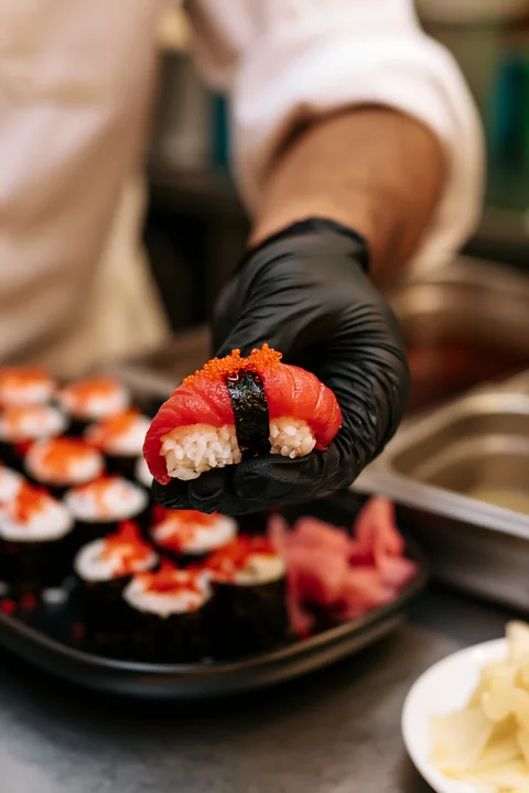 A chef holds one sushi piece with pickled ginger in a busy kitchen.