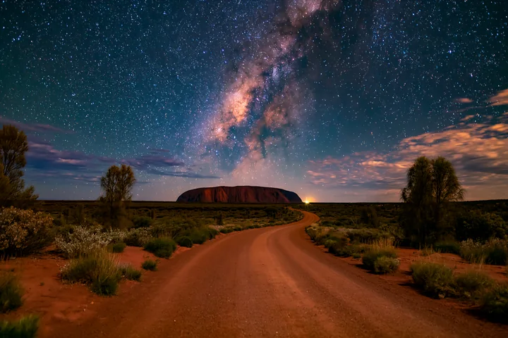 A clear view of Uluru under a starry night sky shows the Milky Way above and a winding dirt road in the foreground.