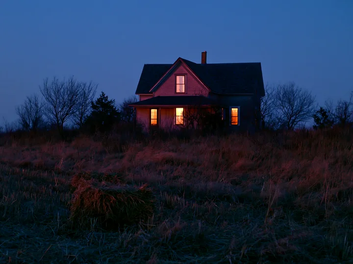 A country home stands alone in a field at night, with light coming from its windows.