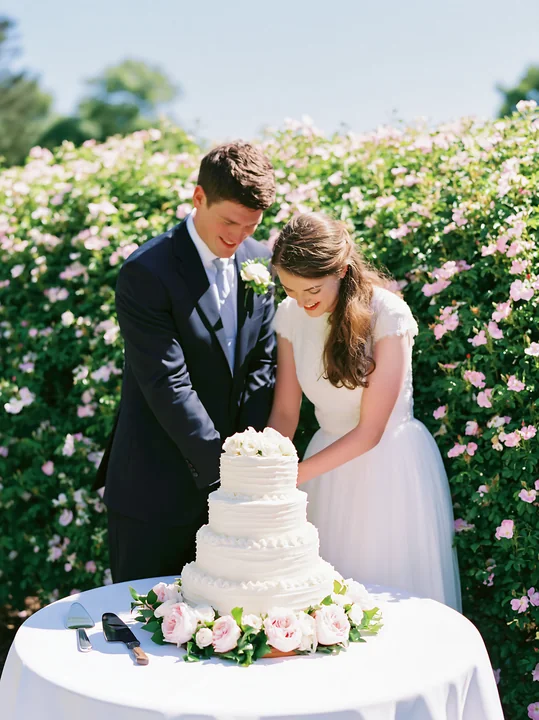 A couple is cutting a wedding cake surrounded by flowers at a garden reception.