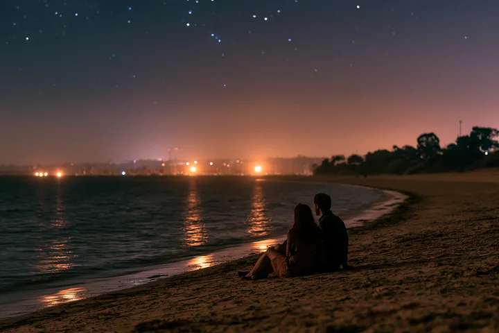 A couple sits on the sand of a beach at night, gazing at the stars above.