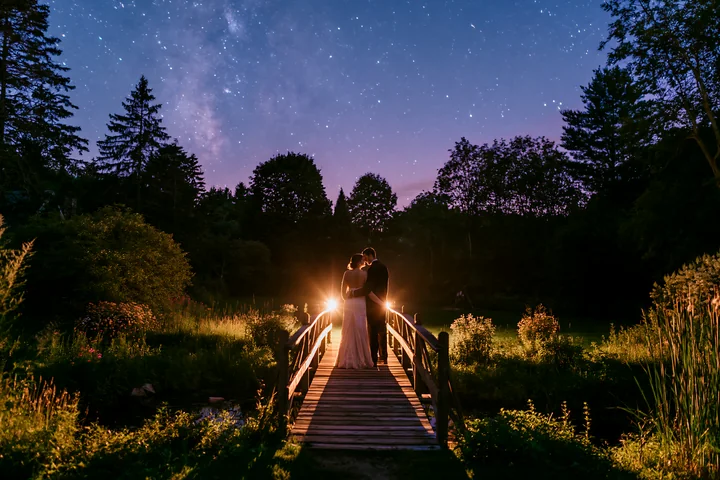 A couple stands on a wooden bridge, sharing a moment under a starry night sky in a rural backyard.