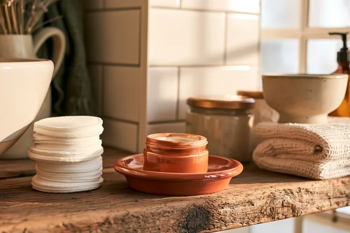 A cream dose beside cotton pads sits on a wooden shelf in a well-lit room.