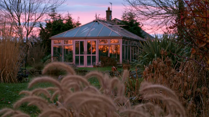 A glass garden conservatory stands surrounded by plants and grass under a colorful evening sky.