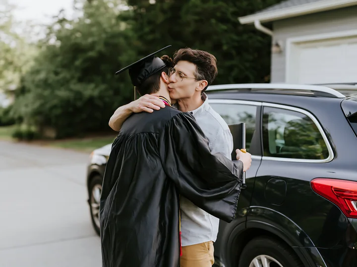 A graduate in a black cap and gown shares a kiss with a loved one in a driveway.
