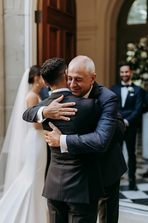 A groom warmly hugs his father during the wedding ceremony inside a grand venue.
