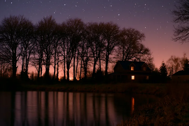 A house stands under stars with silhouette of trees along a calm waterway.