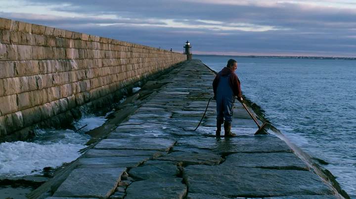 A lighthouse maintenance worker at work stands on a stone pier, using tools to maintain the structure as waves crash nearby.