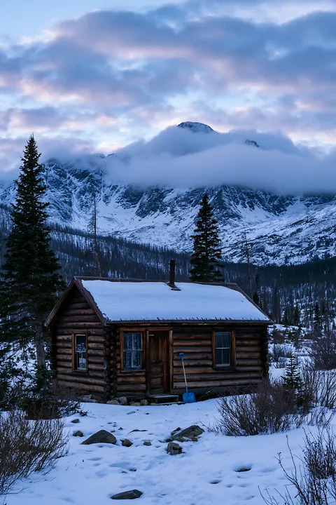 A log cabin stands in the snow with dramatic clouds around alpine peaks in the background.