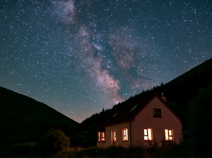 A modern house sits in a rural area with warm lights glowing from its windows as the Milky Way shines brightly overhead.