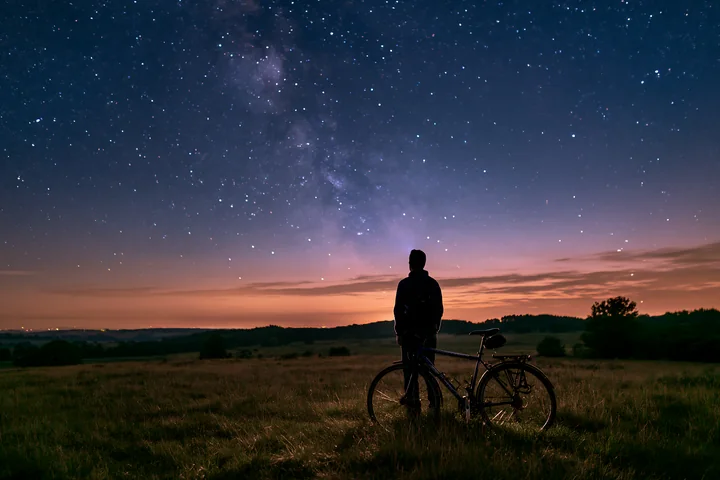 A person stands beside a bicycle, gazing up at a starry sky.
