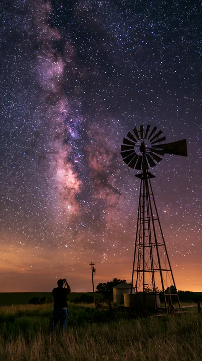 A person using camera under Milky Way stands next to a windmill in a rural setting.