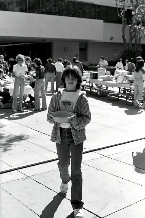 A person walks across an outdoor area holding a bowl at a community potluck buffet.