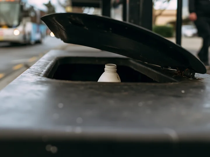 A recycling bin is open, showing a milk bottle placed inside on a busy urban street.