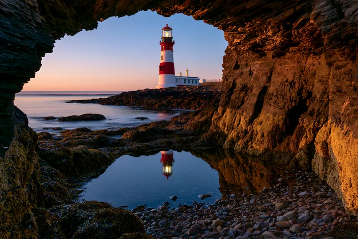 A red and white lighthouse stands near a rocky tide pool during sunset.