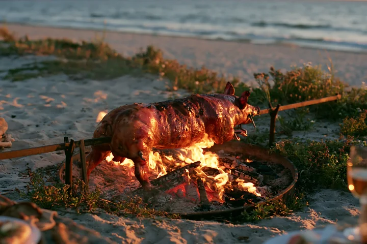 A roasted pig is cooking on a spit over open flames on the beach during sunset.