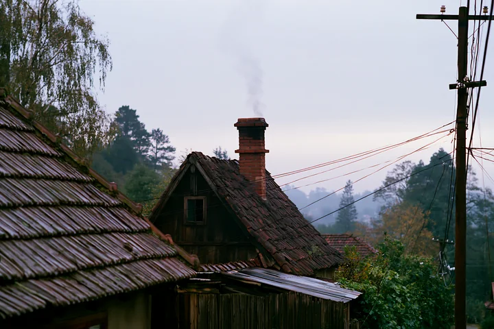 A rural cottage after rain features a chimney with smoke rising, surrounded by trees and power lines.