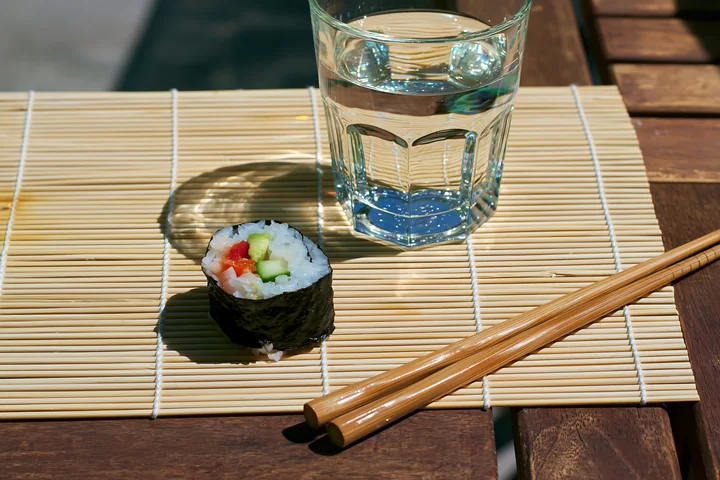 A single sushi roll on a bamboo mat sits next to a glass of water and chopsticks.