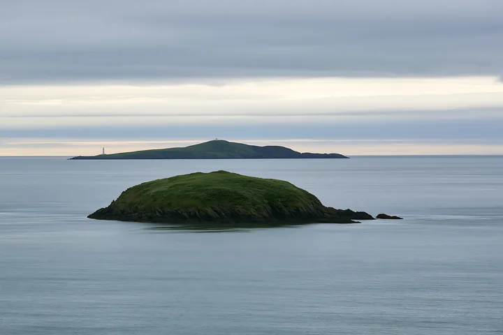A small island with green vegetation is surrounded by water in a calm bay during sunset.