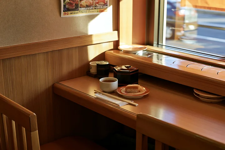 A small table in a sushi conveyor belt restaurant features a plate with sushi, a cup, and condiments.