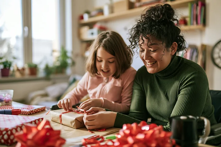 A smiling parent helps a child wrap a gift with paper and ribbon at a home table.