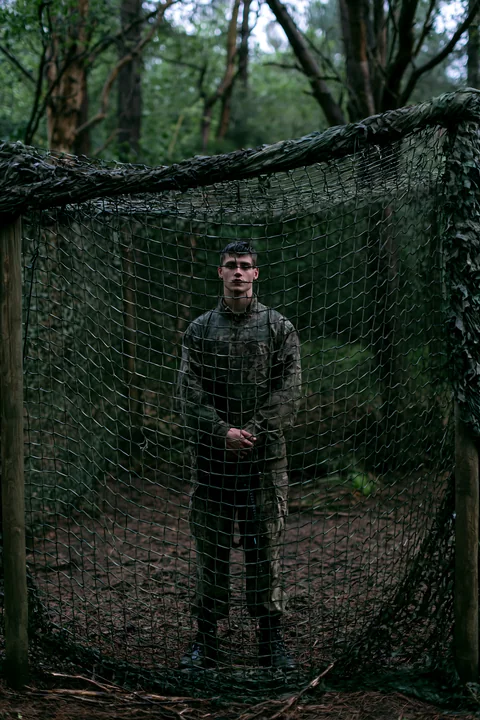 A soldier stands under camouflage netting in a wooded area.