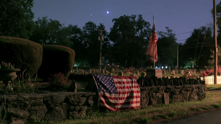 A solemn cemetery scene with American flag and stars shows a flag draped on a stone wall among gravestones under a night sky.