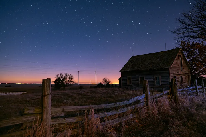 A starry night above a rural wind turbine and a rustic farmhouse shows a clear sky filled with stars.