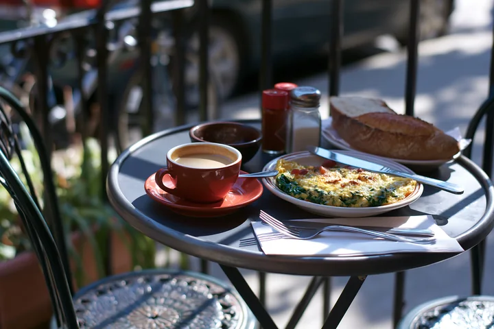A street cafe table holds a cup of milk coffee alongside a plate of breakfast food.