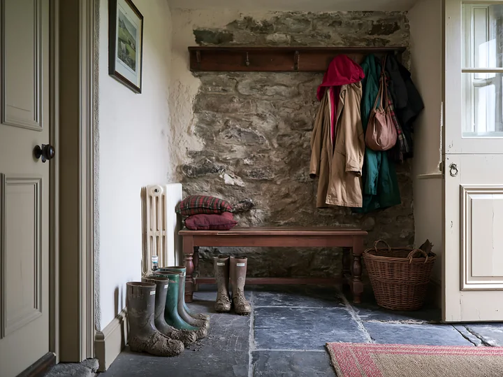 A welcoming farmhouse interior features an exposed stone wall, a wooden bench, and sturdy footwear on a stone floor.