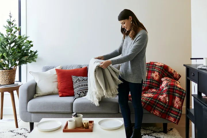 A woman arranges a blanket on a festive sofa with pillows in a modern living room.
