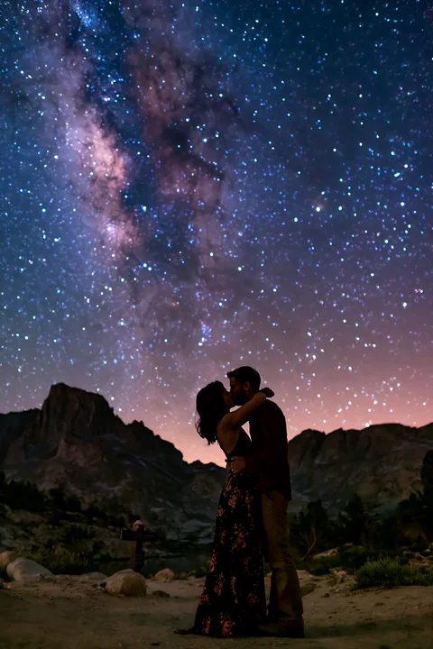A young couple kissing beneath visible Milky Way while surrounded by mountain scenery at night.