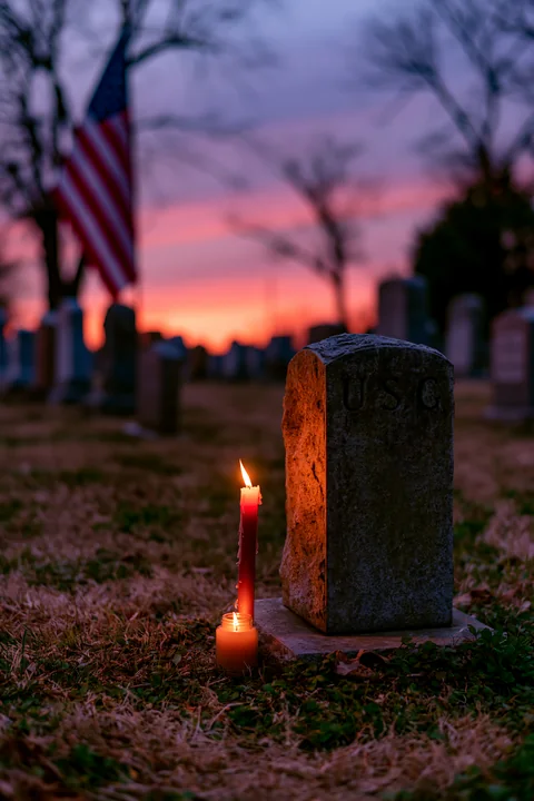 An American flag stands tall above a row of veterans graves as candlelight illuminates the stones during sunset.