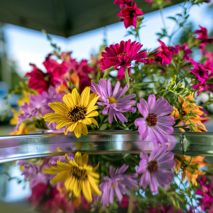 Bright, colorful seasonal flowers are arranged in a glass container at a farmers market.