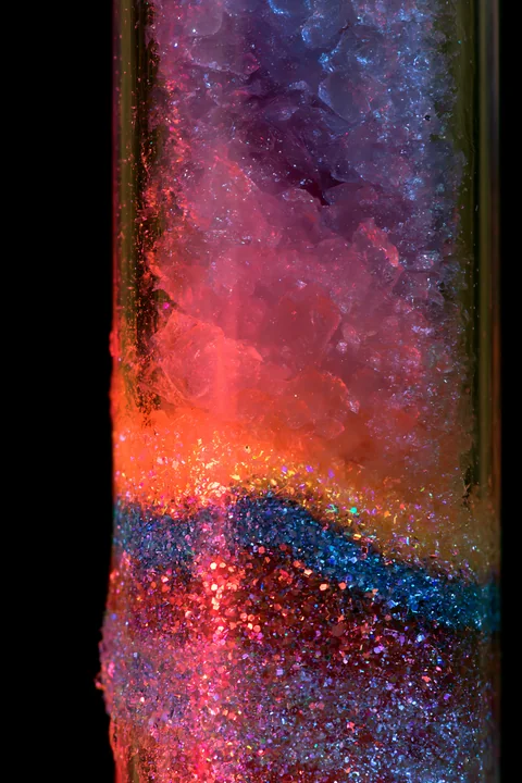 Colorful salt and glitter abstract pattern fills a transparent tube, showcasing layers of blue, pink, and orange materials.