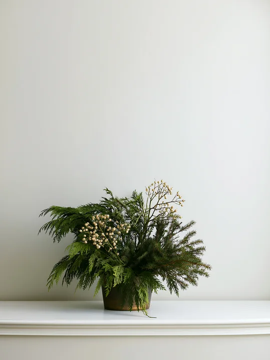 Evergreen Christmas centerpiece with pale berries in a vase on a white mantel, with ample empty wall space above.