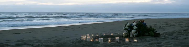 Flowers and lit candles form a memorial on a sandy beach beside the ocean.