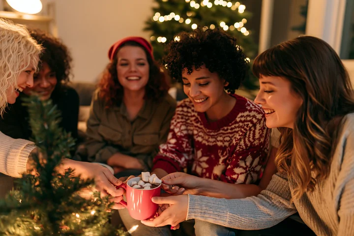 Friends enjoying warm cocoa with marshmallows while celebrating around a Christmas tree with lights in a cozy indoor setting during winter.