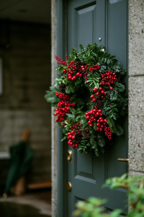 Lush Christmas wreath with red berries and evergreen branches hangs on a dark green apartment door.