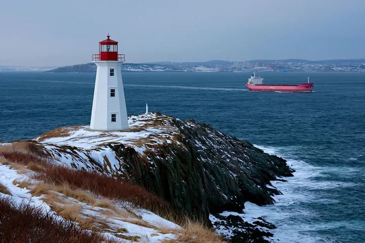 Red-and-white lighthouse on a snowy rocky coast with a cargo ship sailing in the distance.