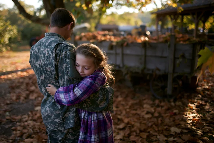 Soldier in camouflage embraces a young child among fallen leaves and rustic outdoor structures.