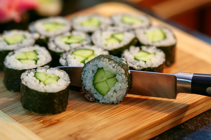 Sushi rolls being cut with knife on a wooden cutting board in a kitchen.