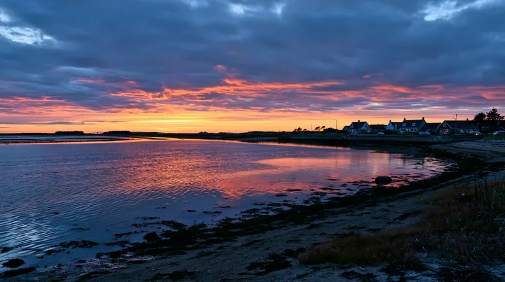 The view shows a coastline with mussel farms at sunset, with reflections in the water and houses along the shoreline.